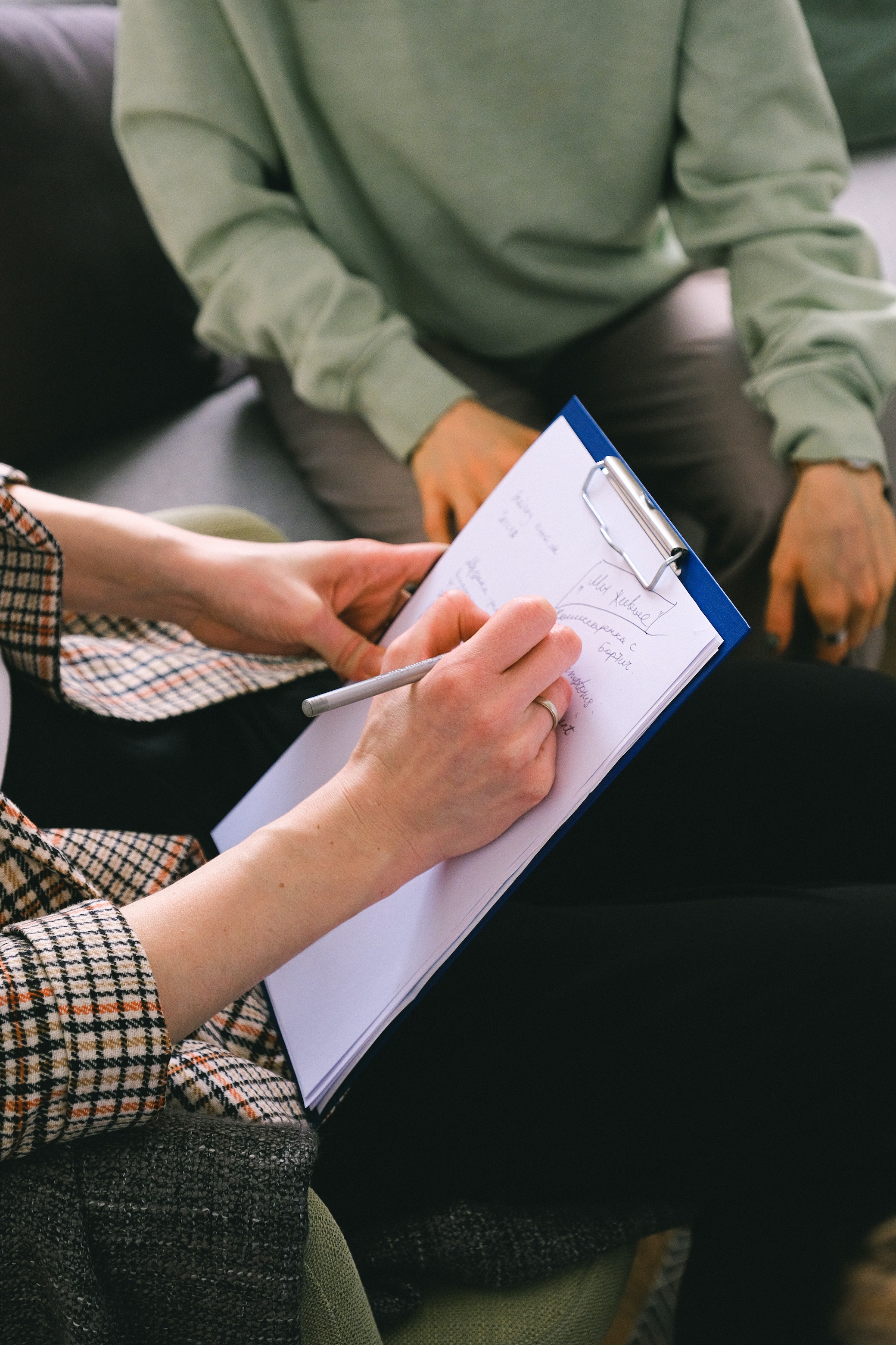 A person holding a clipboard with a form clipped on it, and the person is facing another person sitting on a couch.