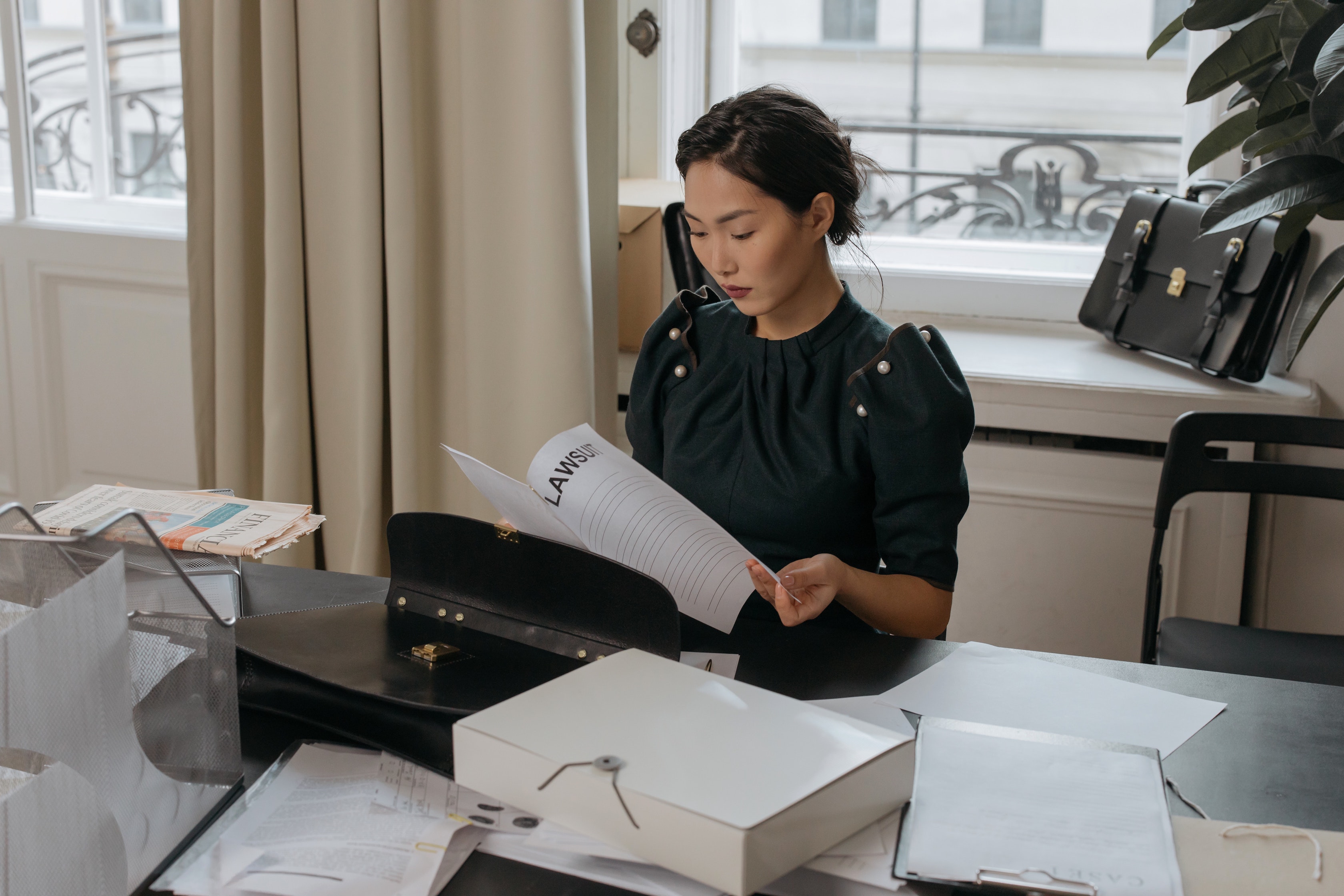 A woman sitting at a desk that has a lot of papers on it. She is looking through some papers that say lawsuit on the first page.